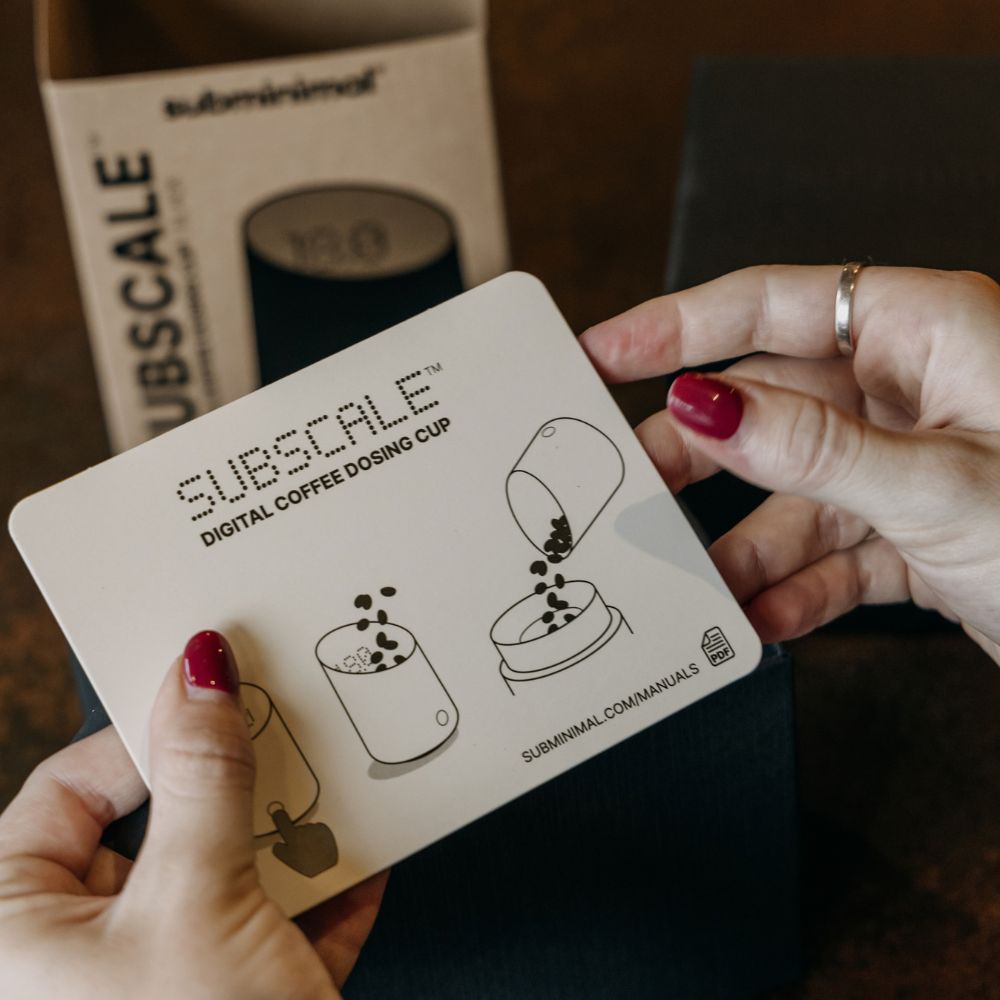 A person's hands holding a white Subscale digital coffee dosing cup instruction card, with the Subscale box in the background on a dark wooden surface.