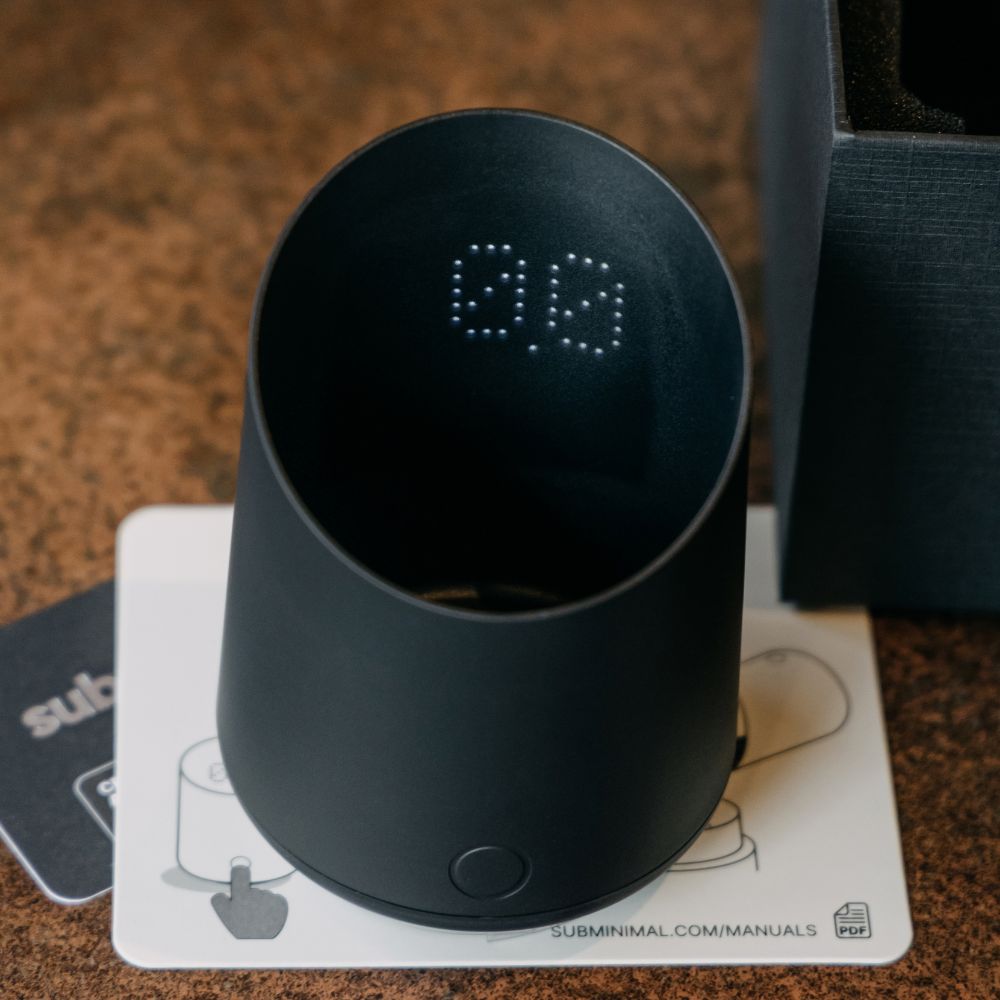 A matte black cylindrical smart speaker or device with a digital LED display showing '00' inside the open top, resting on a white instruction card and next to its packaging box on a brown textured surface.